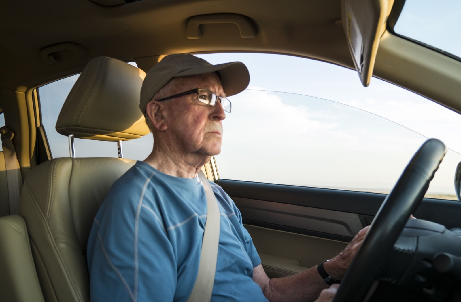 An older adult man wearing glasses driving with the car window beside him partially opened.