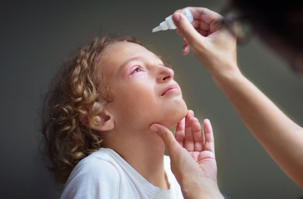 A child with pink eye is assisted by an adult with eye drops.