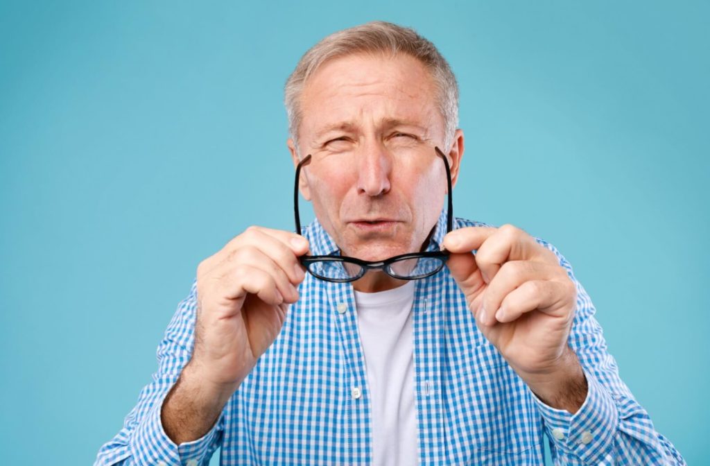 A senior man holding eyeglasses with both hands and squinting from blurry vision.