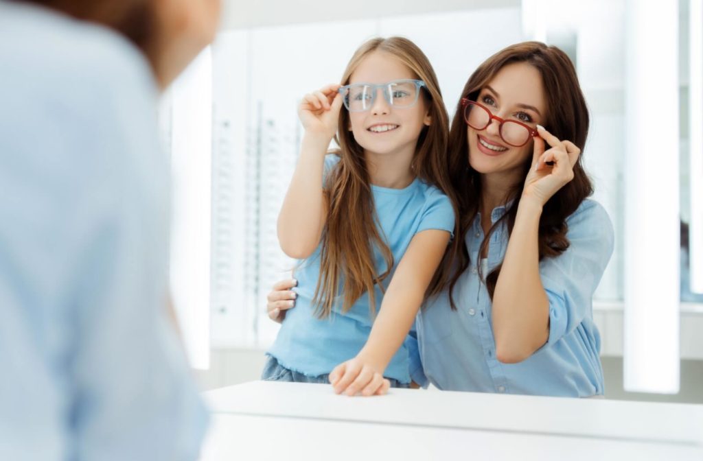 A parent and child smile at themselves in the mirror of an eye doctor’s office as they try on eyeglass frames