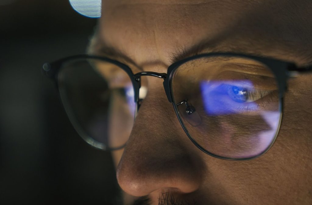 A close-up image of a person wearing glasses. The light of their computer screen is reflected on their lenses.