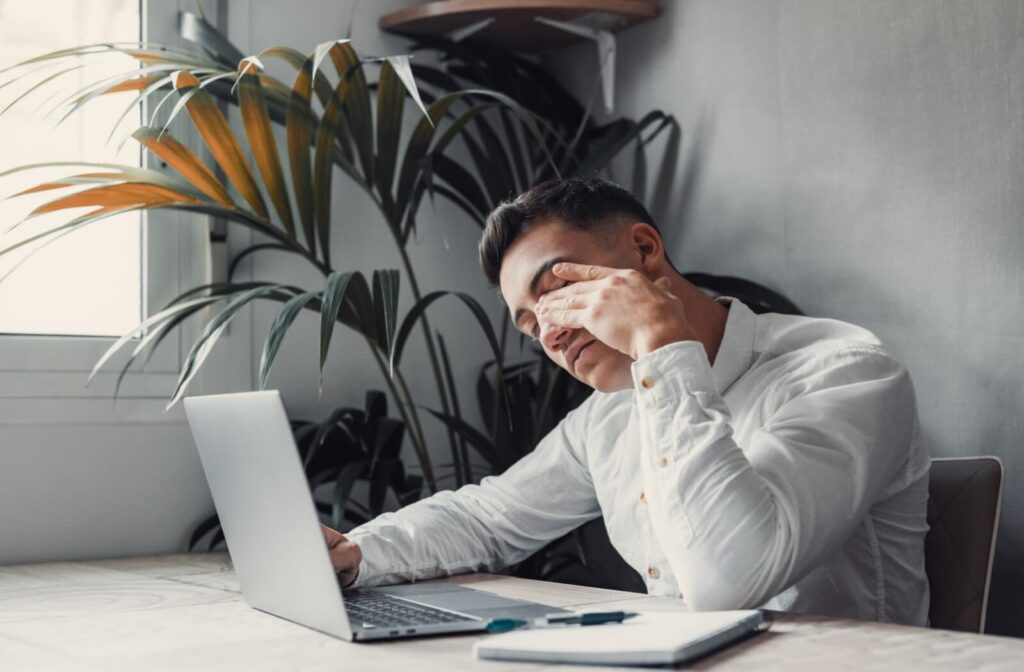 A young adult rubbing their eyes in frustration due to astigmatism vision problems while working from home.