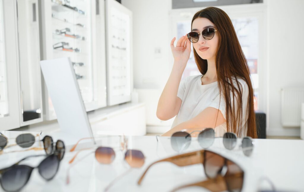 A fashionable young adult tries on different sunglasses while looking in a mirror at their optometrist's office.