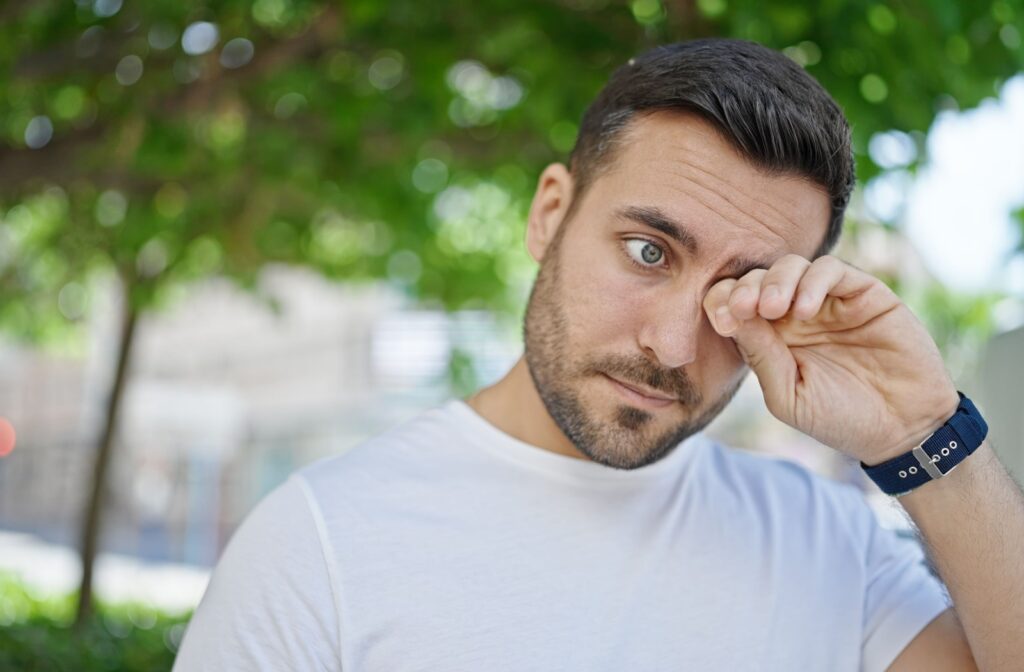 A person wearing a white shirt rubs their eye while standing outdoors under green trees.