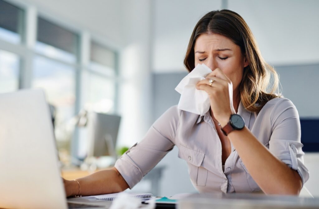A person sitting a computer working and blowing their nose due to seasonal allergies.