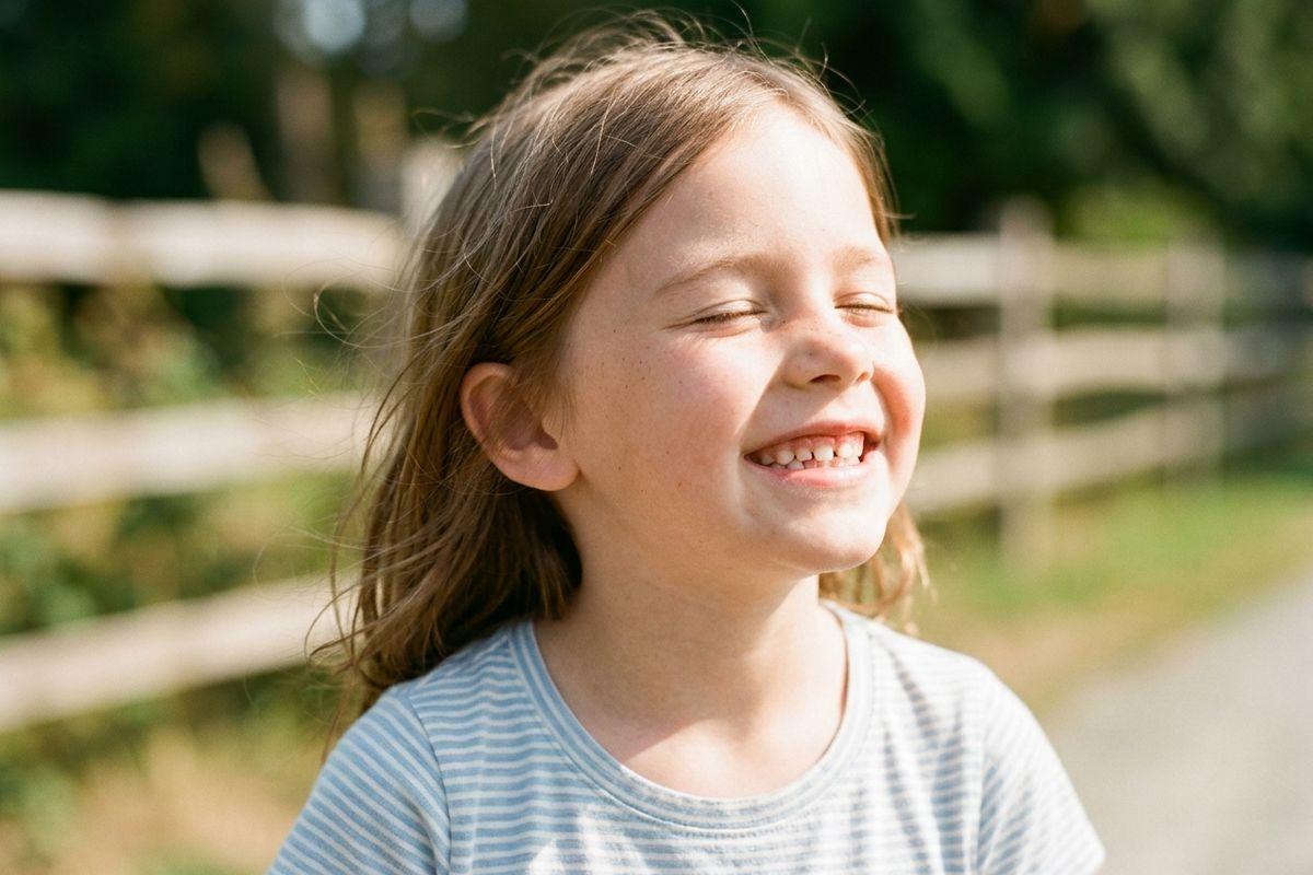 A child with long brown hair laughing outdoors near a wooden fence on a sunny day, eyes closed, wearing a striped shirt.